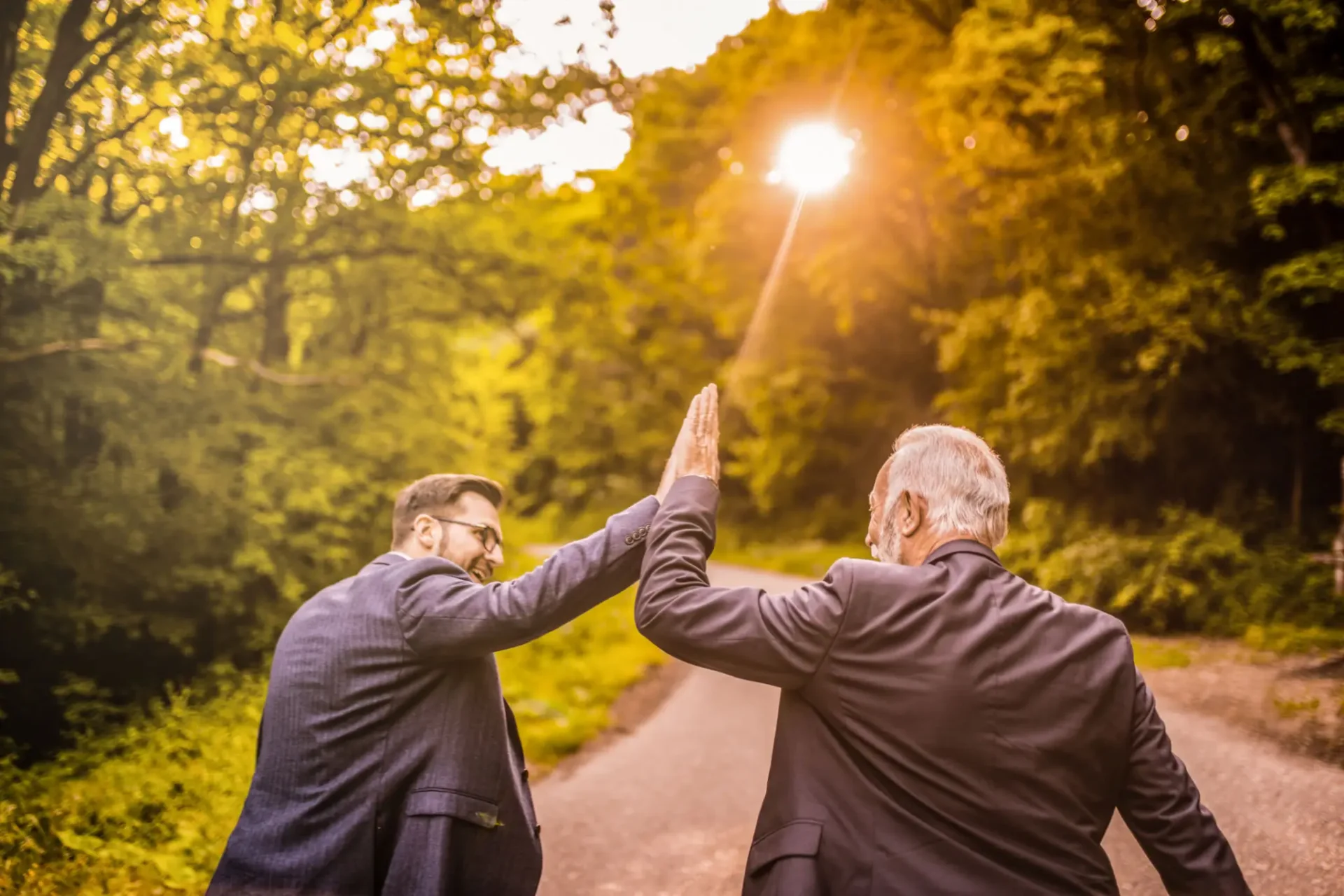 Two men in suits high-fiving on sunlit path